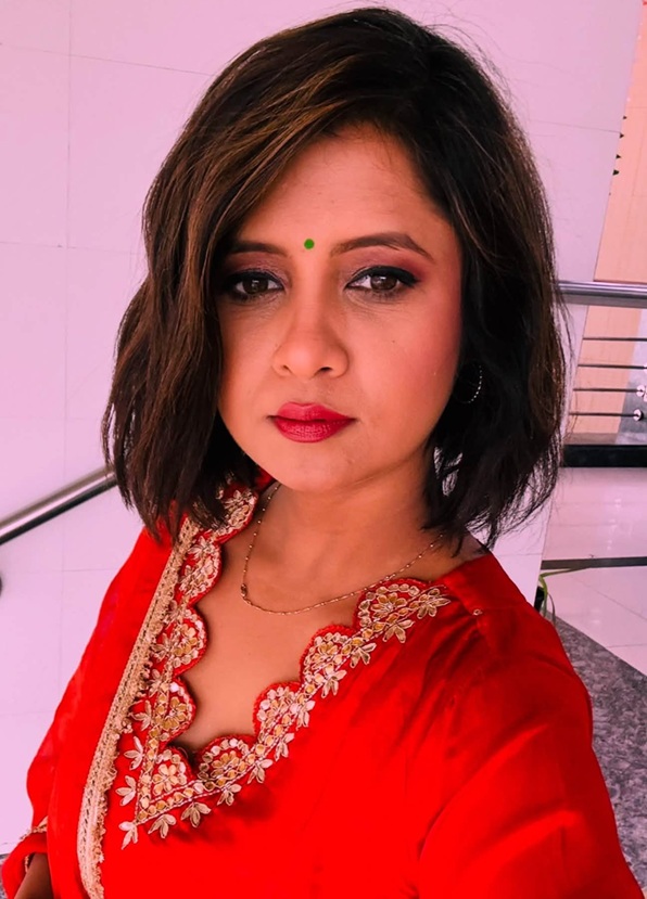 Portrait of a woman with short dark hair wearing a red traditional top with gold floral embroidery and a green bindi looking at the camera.