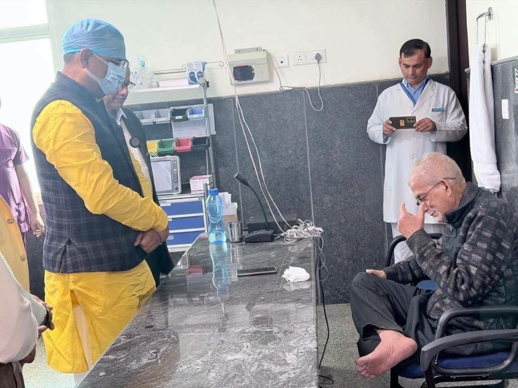 Medical team in a clinic room shows debriefing a seated elderly patient; one doctor in yellow suit and mask stands near a glass desk, another in white coat watches on the wall.