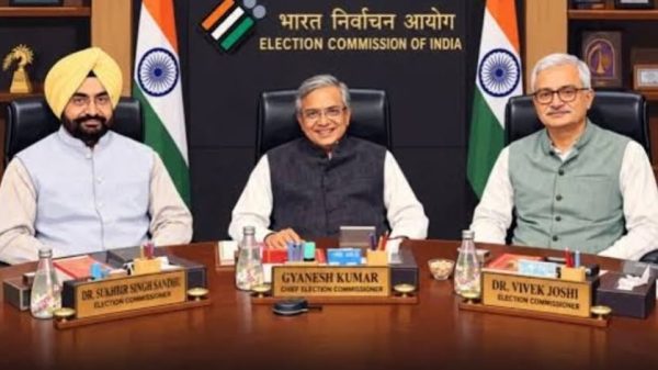 Three men sit at a conference table in the Election Commission of India office, with Gyanesh Kumar, Chief Election Commissioner, centered; Indian flags and the EC backdrop behind them.