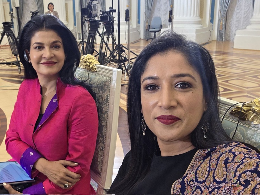 Two women pose for a selfie at a formal event, with cameras and a grand room visible in the background.