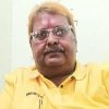 Man with glasses and mustache wearing a yellow polo shirt indoors, with a tilak on his forehead.
