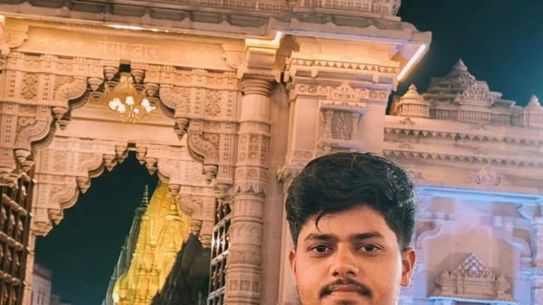 Young man in a pink shirt stands in front of an ornate temple gateway lit at night, with intricate carvings surrounding him.
