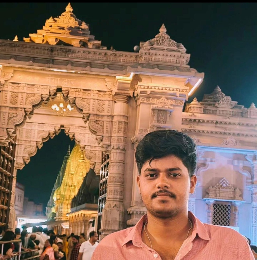 Young man in a pink shirt stands in front of an ornate temple gateway lit at night, with intricate carvings surrounding him.