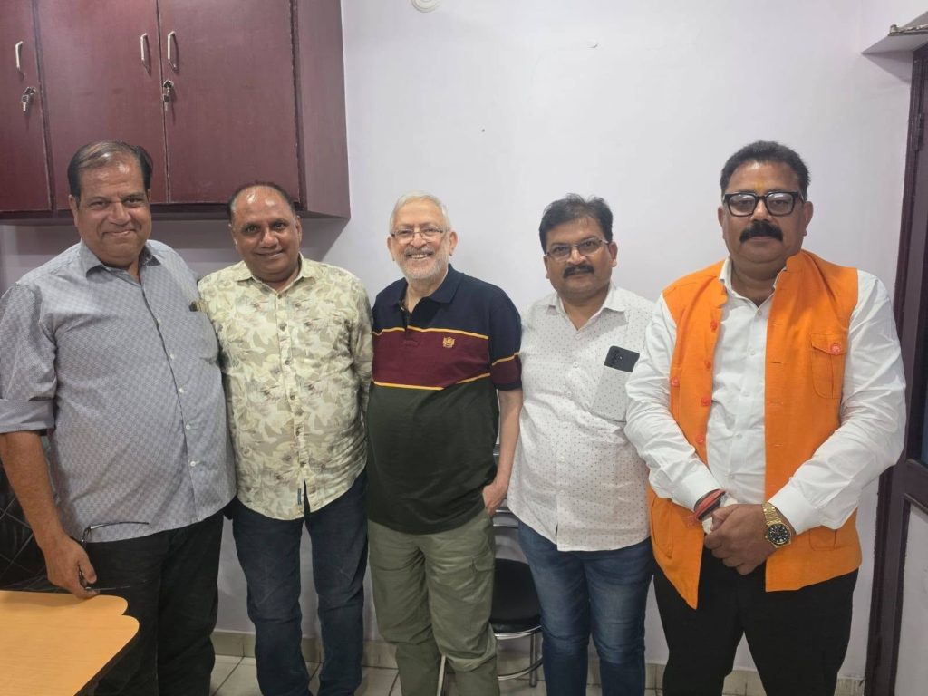 Five men stand side by side indoors, smiling at the camera; one wears an orange vest over a white shirt, others in casual shirts.