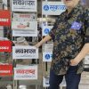 Older man wearing glasses and a floral shirt stands beside a newspaper rack displaying Navbharat and Sakal logos.