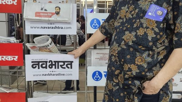 Older man wearing glasses and a floral shirt stands beside a newspaper rack displaying Navbharat and Sakal logos.