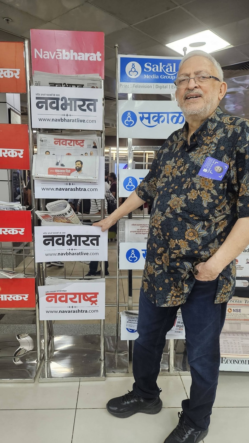 Older man wearing glasses and a floral shirt stands beside a newspaper rack displaying Navbharat and Sakal logos.