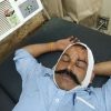 Man with a bandaged head and stitched wound rests on a hospital bed, eyes closed, wearing a blue striped shirt.
