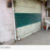 Closed storefront with a green striped metal shutter and a white plastic chair on the sidewalk beside an outdoor AC unit on the wall edge of a building.