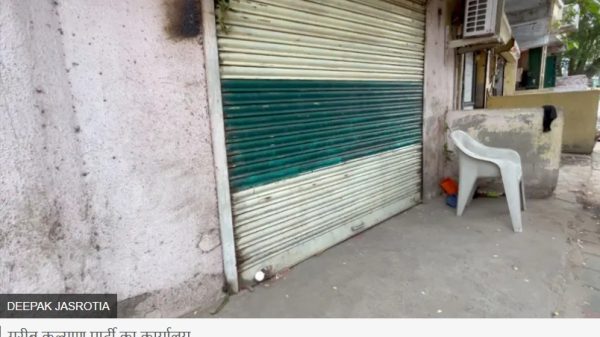 Closed storefront with a green striped metal shutter and a white plastic chair on the sidewalk beside an outdoor AC unit on the wall edge of a building.