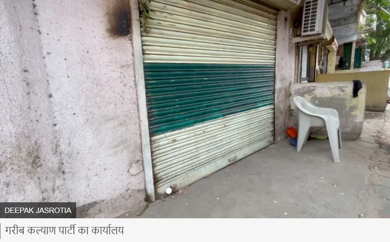 Closed storefront with a green striped metal shutter and a white plastic chair on the sidewalk beside an outdoor AC unit on the wall edge of a building.