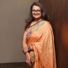Smiling woman wearing an orange embroidered saree and glasses, standing against a wooden wall.