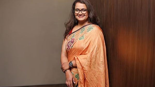 Smiling woman wearing an orange embroidered saree and glasses, standing against a wooden wall.