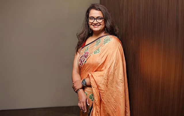 Smiling woman wearing an orange embroidered saree and glasses, standing against a wooden wall.