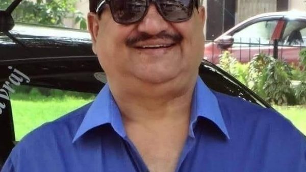 Smiling man with dark hair and sunglasses wearing a blue button-down shirt outdoors, with a car and greenery in the background.