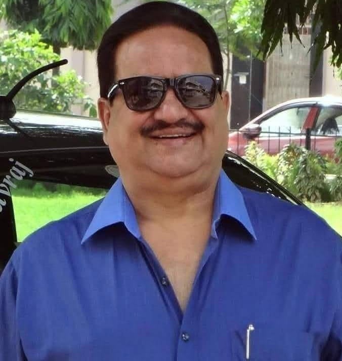 Smiling man with dark hair and sunglasses wearing a blue button-down shirt outdoors, with a car and greenery in the background.