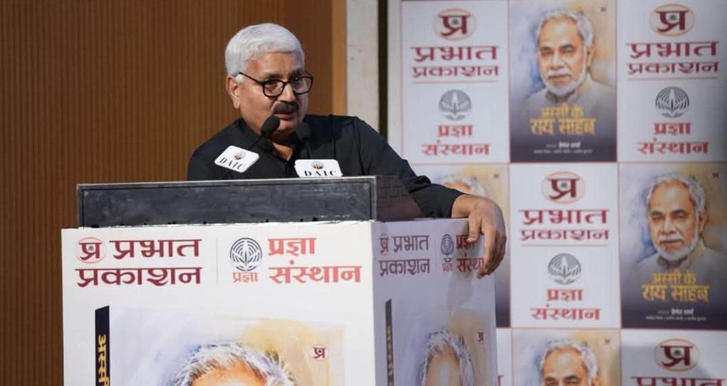 hemant sharma | No. 1 Indian Media News Portal Man with glasses and gray hair speaks at a podium with microphones, at a backdrop covered in posters featuring Hindi text and a portrait.