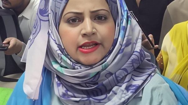 Woman wearing a patterned hijab speaks into a microphone labeled DigiFirst, with reporters behind her in a crowded indoor setting.