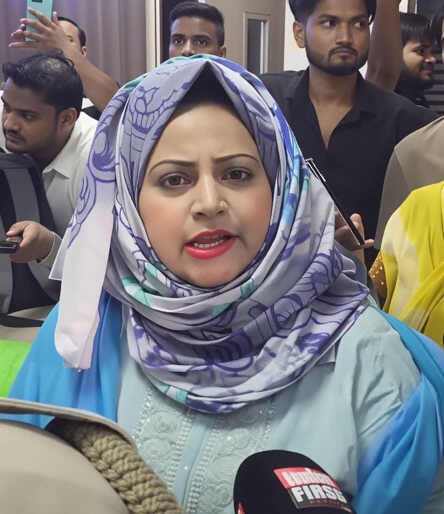 Woman wearing a patterned hijab speaks into a microphone labeled DigiFirst, with reporters behind her in a crowded indoor setting.