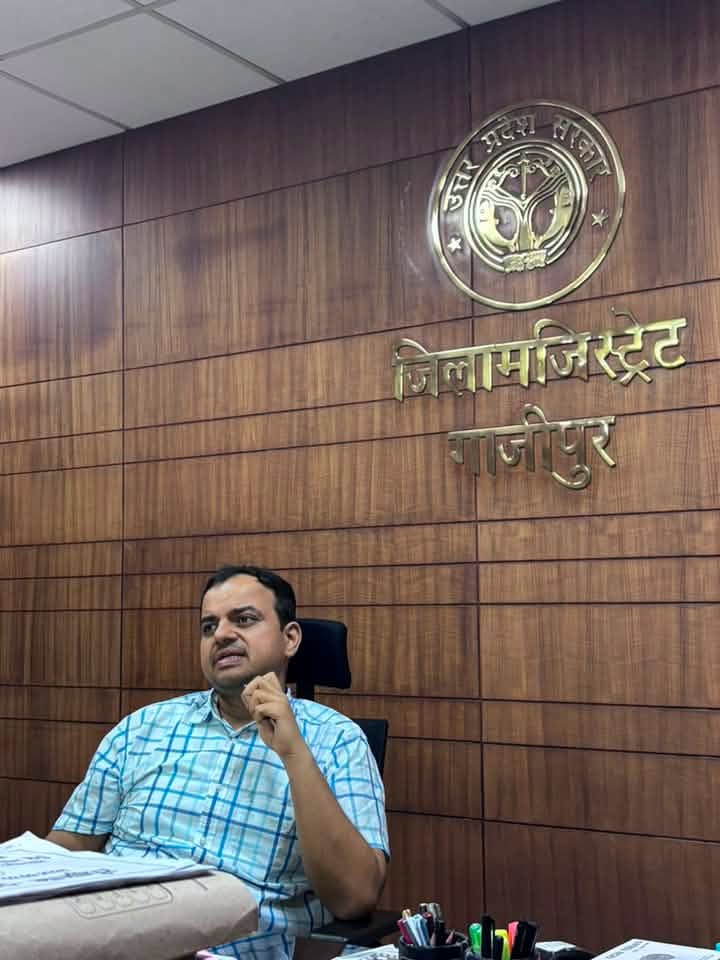 ias anupam shukla | No. 1 Indian Media News Portal Man in a blue checked shirt sits at a desk in a government office, wood-paneled wall behind with a circular seal and Hindi signage.