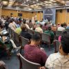 Audience in a lecture hall watching a speaker at a podium with a blue banner on stage.