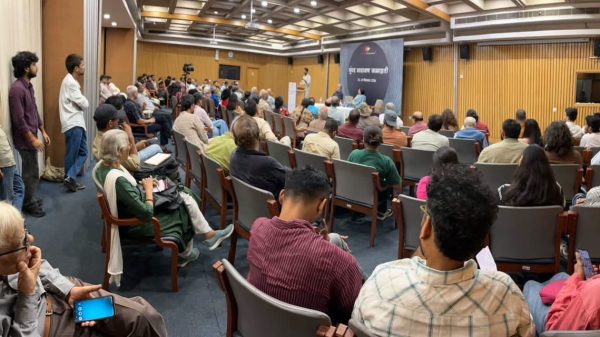 Audience in a lecture hall watching a speaker at a podium with a blue banner on stage.