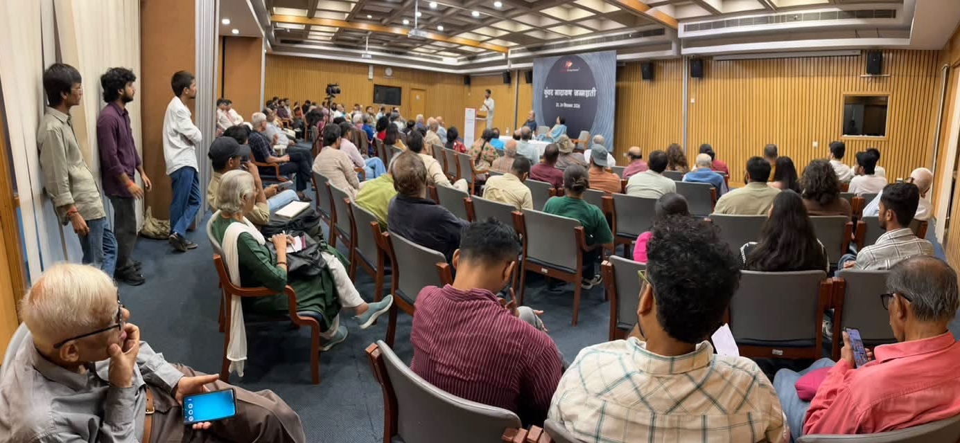 Audience in a lecture hall watching a speaker at a podium with a blue banner on stage.