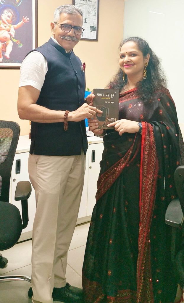 img_0825 | No. 1 Indian Media News Portal Man in a dark waistcoat hands a book to a woman in a black saree with a red border in an office, both smiling.