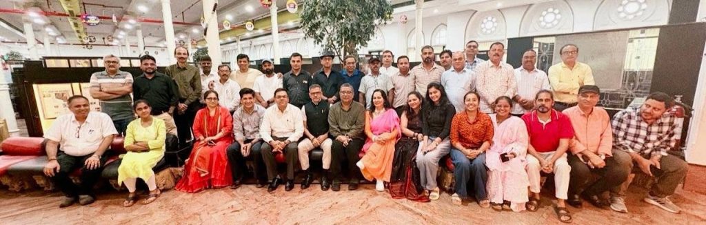img_0828 | No. 1 Indian Media News Portal Group photo of a diverse crowd of adults posing together in a bright indoor lobby or mall setting, seated and standing in three rows.