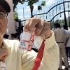 Man wearing a cap and glasses holds up red ID lanyards outside a gated fence; security officers are in the background.