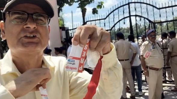 Man wearing a cap and glasses holds up red ID lanyards outside a gated fence; security officers are in the background.