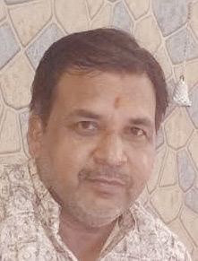 Portrait of a man with short hair and a mustache, wearing a light patterned shirt, with a tilak on his forehead, in front of a patterned wall.