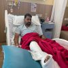 Man in a hospital bed with a red blanket and a leg cast, looking at the camera in a clinical room.