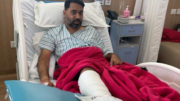 Man in a hospital bed with a red blanket and a leg cast, looking at the camera in a clinical room.