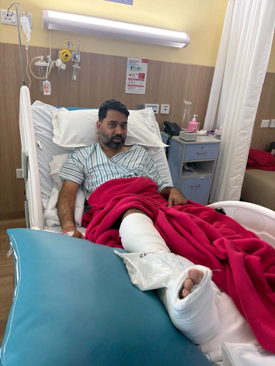 Man in a hospital bed with a red blanket and a leg cast, looking at the camera in a clinical room.