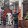 People standing on a rainy street by a gate; a man in an orange turban sprays orange paint on a wall while others hold umbrellas.