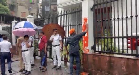 People standing on a rainy street by a gate; a man in an orange turban sprays orange paint on a wall while others hold umbrellas.