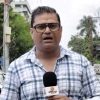 Male news reporter in glasses and a blue plaid shirt holds a microphone, reporting outdoors on a street with trees in the background.