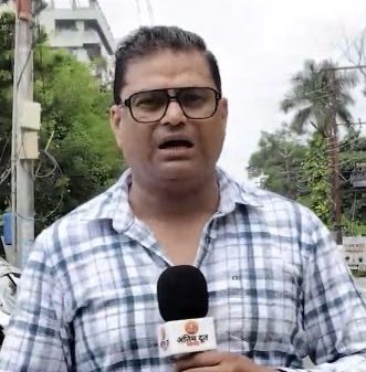 Male news reporter in glasses and a blue plaid shirt holds a microphone, reporting outdoors on a street with trees in the background.