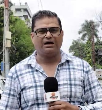 Male news reporter in glasses and a blue plaid shirt holds a microphone, reporting outdoors on a street with trees in the background.