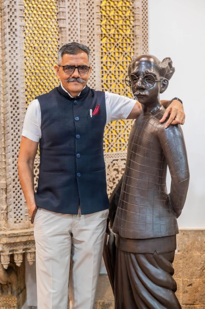 img_0889 | No. 1 Indian Media News Portal A man in a navy blue vest with a white shirt stands beside a bronze statue wearing round glasses in a decorative setting.