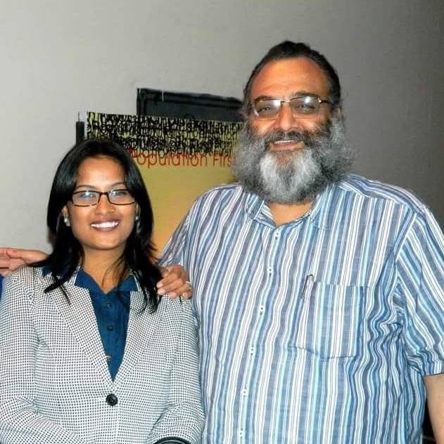 Woman in a blue blouse and checkered blazer stands next to a bearded man in a blue striped shirt; both smile at the camera with a poster in the background.