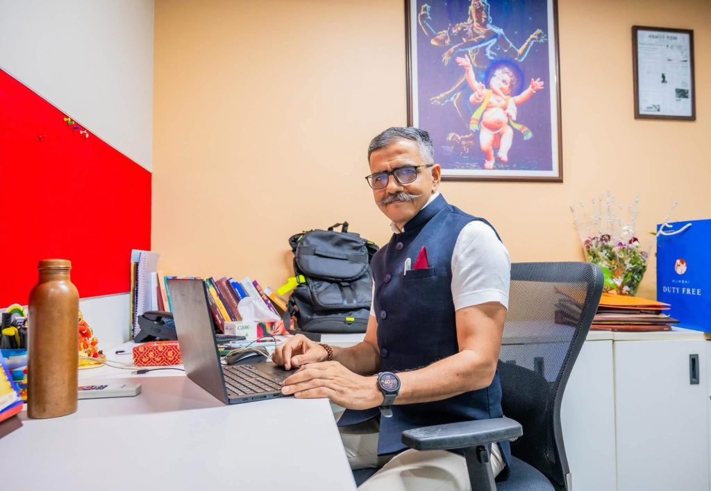 Man in a vest and glasses sits at a desk typing on a laptop in a colorful office.