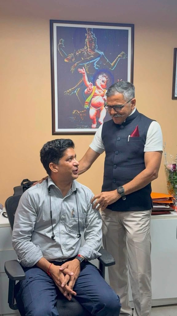 Two men in an office: a seated doctor wearing a light shirt with a stethoscope, and a standing colleague in a dark vest smiling beside him.
