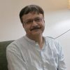 Man with glasses and a mustache, wearing a light striped shirt, seated on a sofa indoors and smiling slightly.