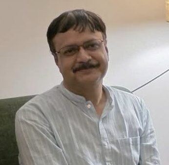 Man with glasses and a mustache, wearing a light striped shirt, seated on a sofa indoors and smiling slightly.