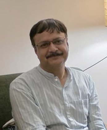 Man with glasses and a mustache, wearing a light striped shirt, seated on a sofa indoors and smiling slightly.
