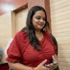 Smiling woman in a red blouse walks through a wood-paneled hallway while holding a smartphone.