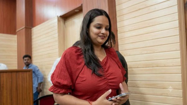 Smiling woman in a red blouse walks through a wood-paneled hallway while holding a smartphone.