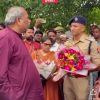 Man in a maroon kurta hands a bouquet to a uniformed police officer as a crowd looks on during an outdoor event.